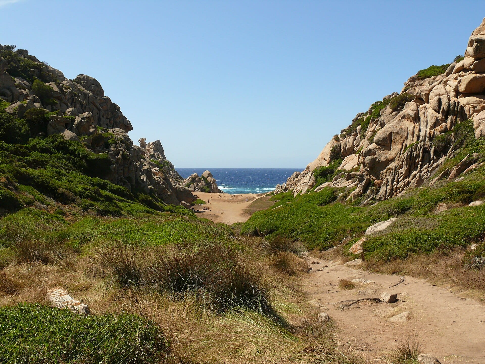 Blick auf das Valle della Luna, Capo Testa, mit schroffen Granitfelsen, grünen Büschen, sandigem Talboden und dem blauen Mittelmeer im Hintergrund.