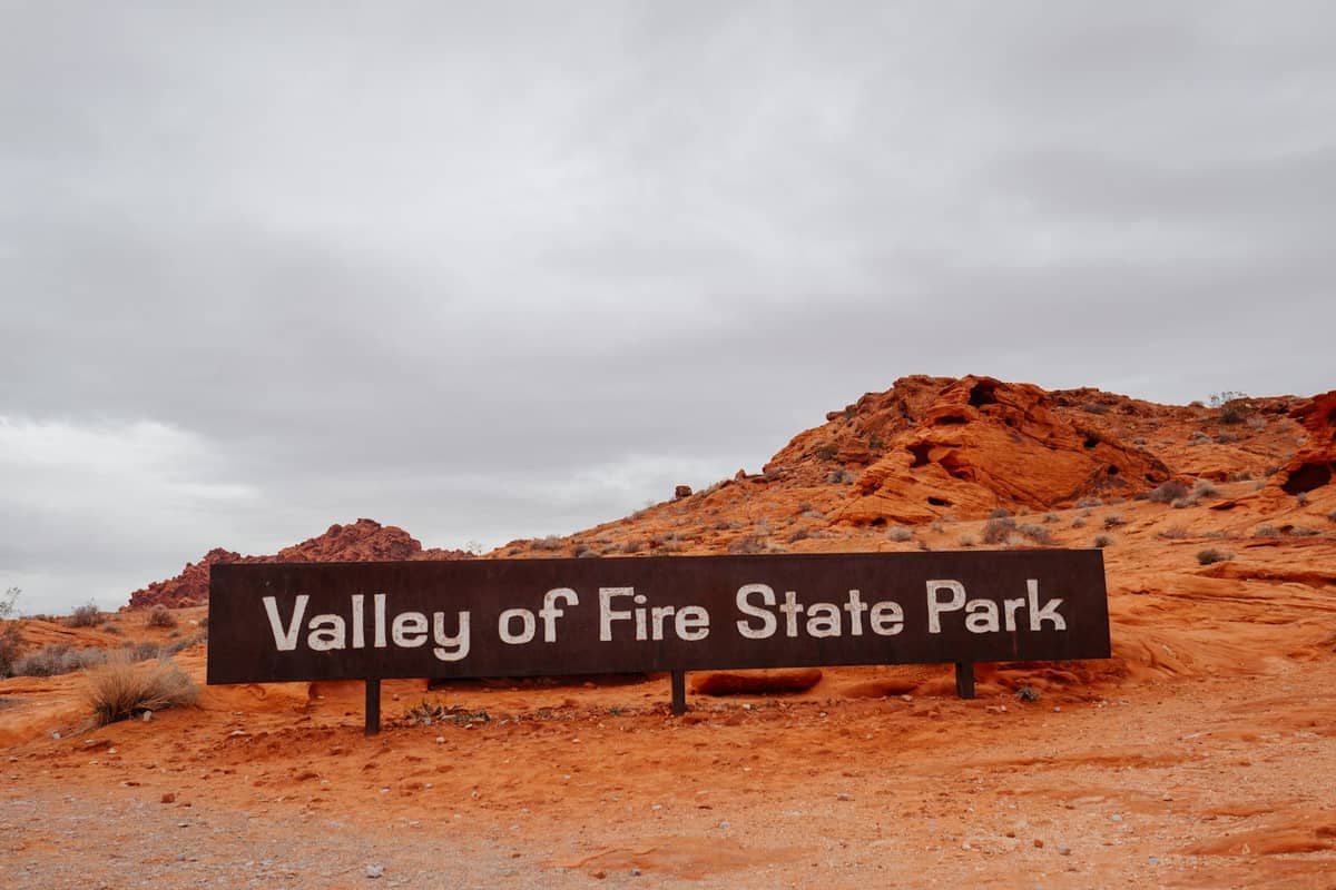 Papan tanda masuk Valley of Fire State Park dikelilingi formasi batu pasir merah yang cerah dan langit gurun berawan.