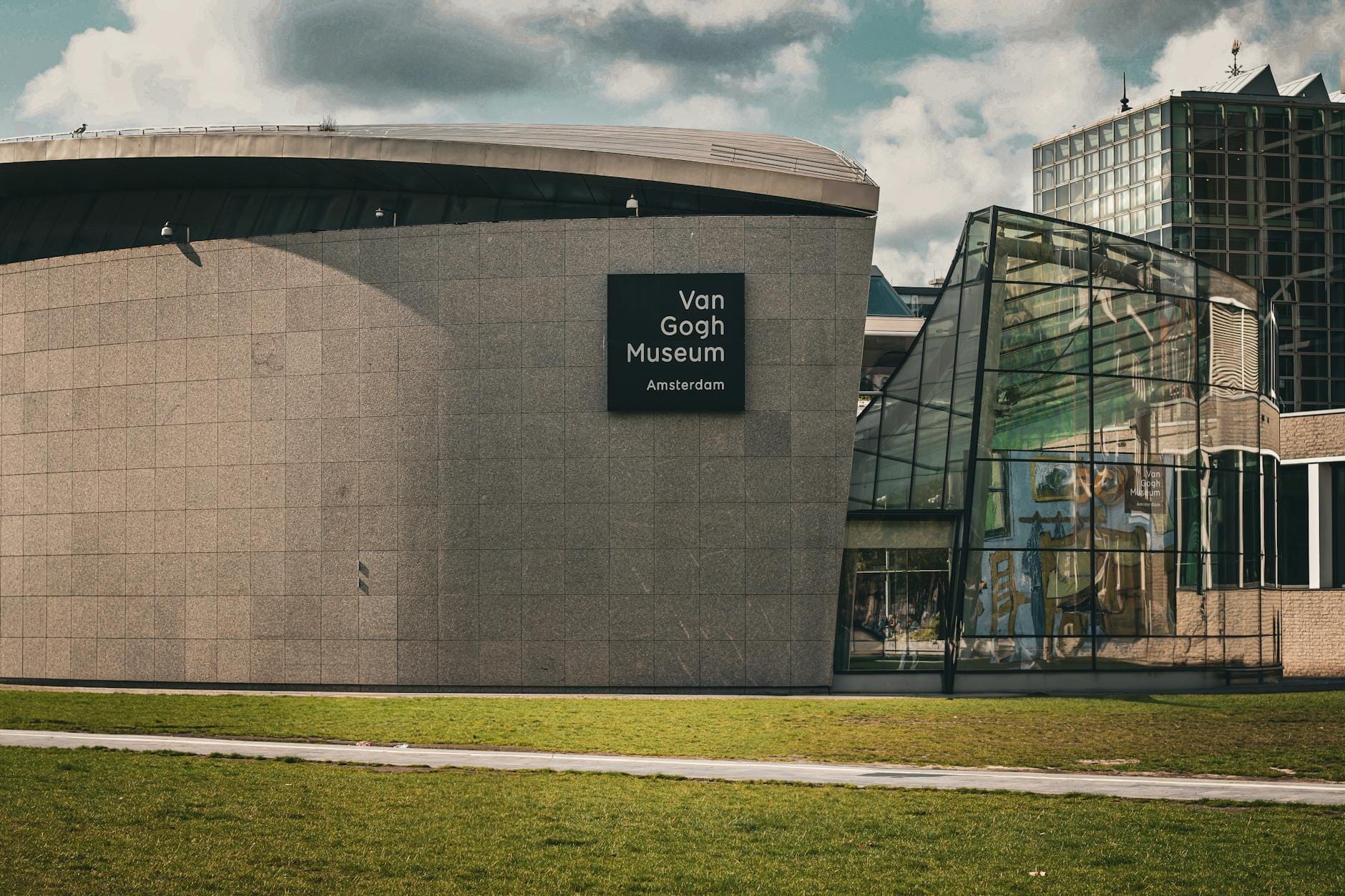 El exterior del Museo Van Gogh en Ámsterdam, con su característica arquitectura moderna, la entrada principal y el césped verde bajo un cielo parcialmente nublado.