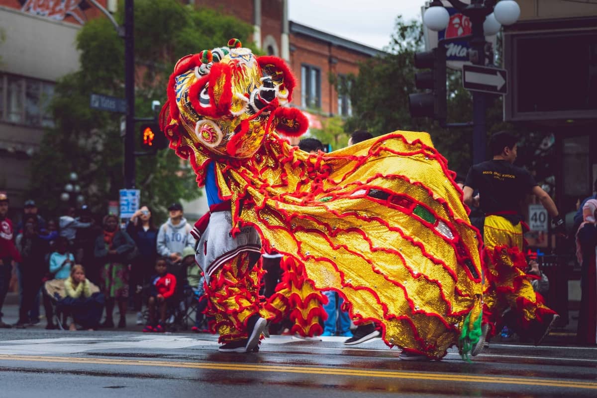 Ein leuchtend gelbes und rotes Löwentanz-Kostüm, das während einer Parade auf einer Stadtstraße aufgeführt wird, mit Zuschauern und Chinatown-Gebäuden im Hintergrund.