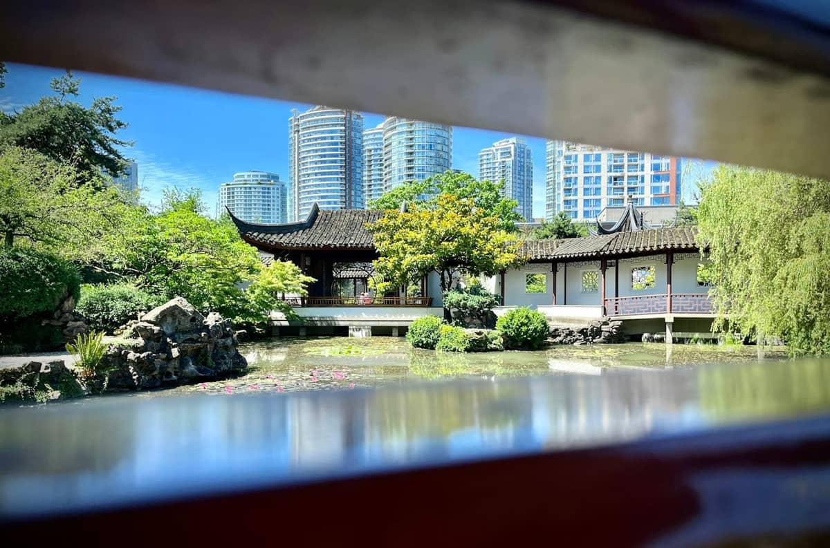 Klassischer chinesischer Garten mit verziertem Pavillon, üppigem Grün und der Skyline von Vancouver im Hintergrund unter blauem Himmel.