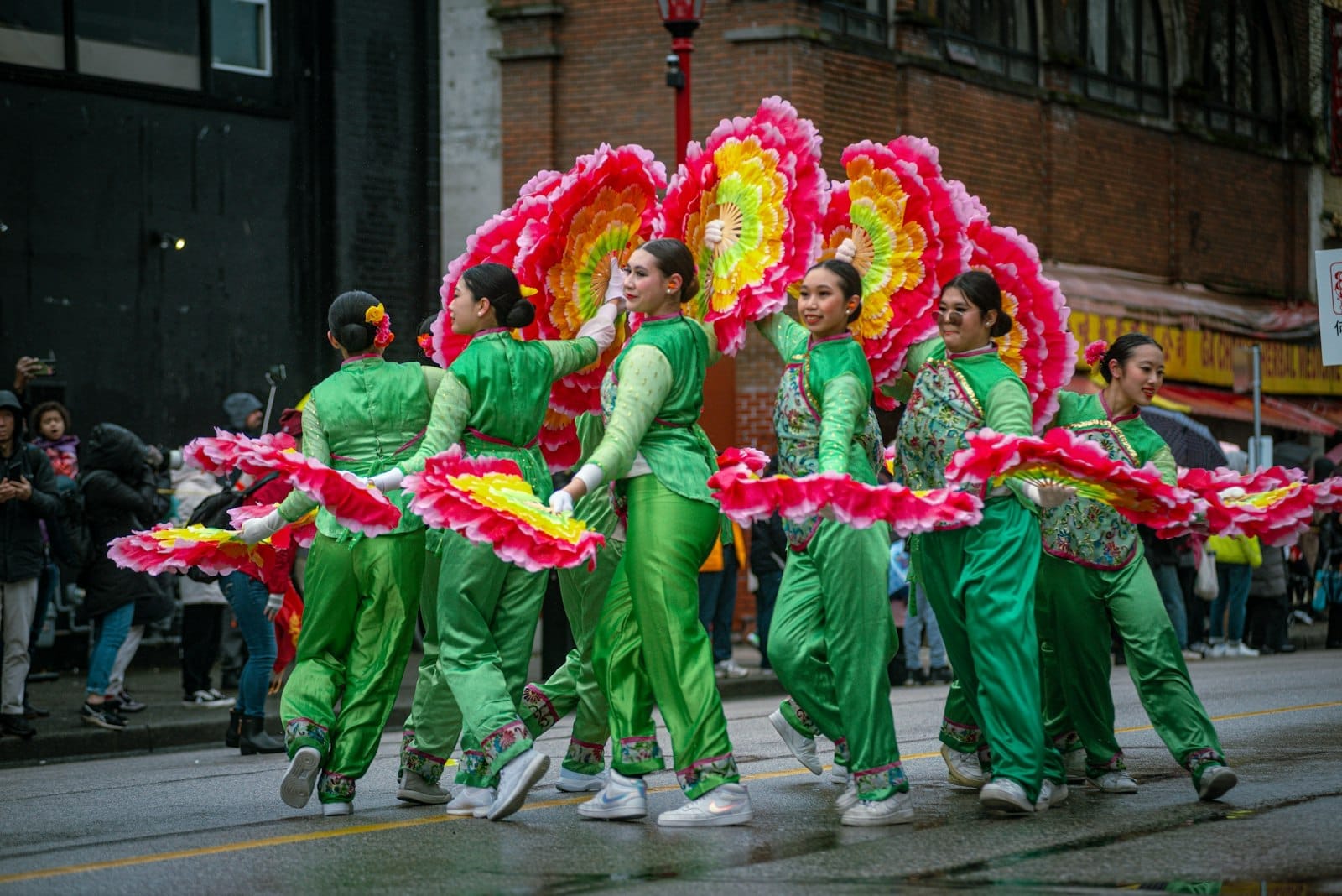 Grupo de mulheres em trajes verdes vibrantes dançando em um desfile tradicional com grandes leques de flores rosa e amarelas em uma rua chuvosa no bairro chinês de Vancouver.