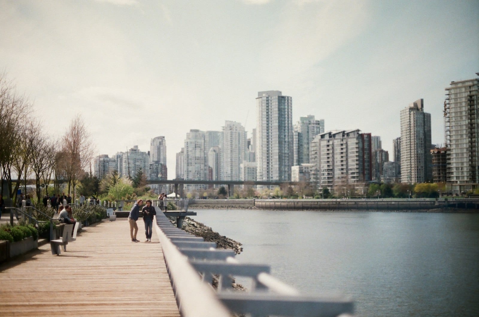 Casal de mãos dadas caminhando em um calçadão de madeira à beira-mar com o skyline de Vancouver ao fundo em um dia ensolarado.