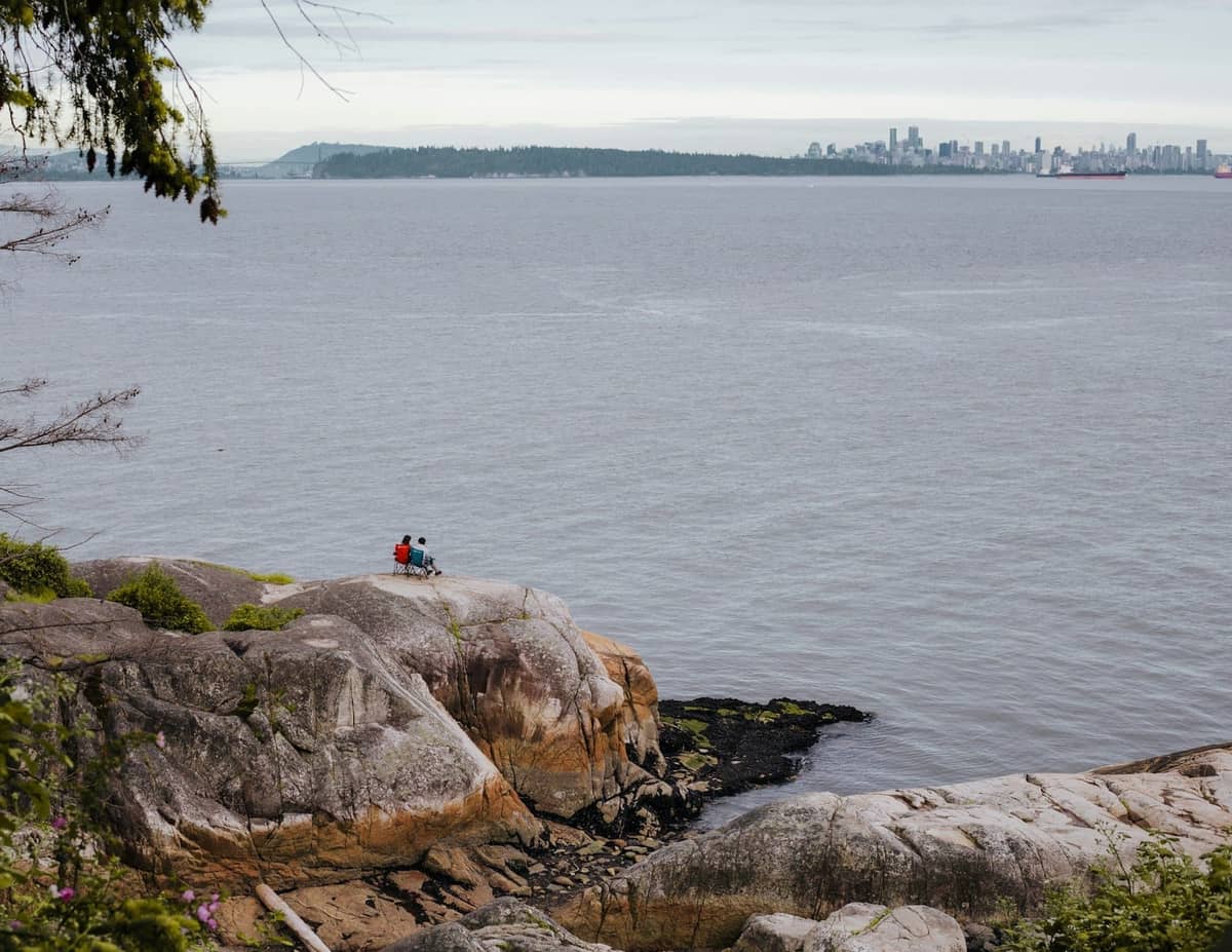 Paar sitzt auf einem Felsvorsprung mit Blick auf den Ozean und die Skyline von Vancouver in der Ferne, umgeben von natürlicher Küstenlandschaft.