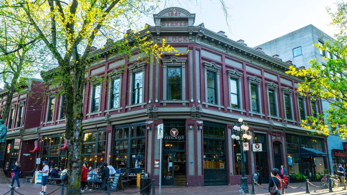 Historisches Backsteingebäude mit großen Fenstern und Restaurants an einer baumgesäumten Ecke im Gastown-Viertel von Vancouver