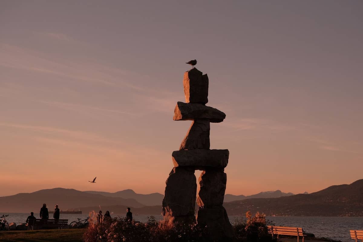 Inukshuk-Steinstatue nahe der Uferpromenade von Vancouver bei Sonnenuntergang, mit Bänken, Wasser und Bergen im Hintergrund.