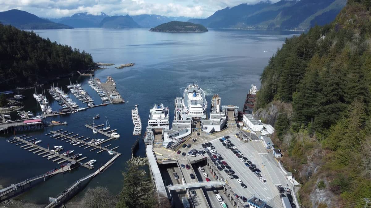 Luftaufnahme eines großen Fährterminals in Vancouver mit mehreren angedockten BC Ferries-Schiffen, Fahrspuren für Fahrzeuge, Marina und malerischen Küstengebirgen.