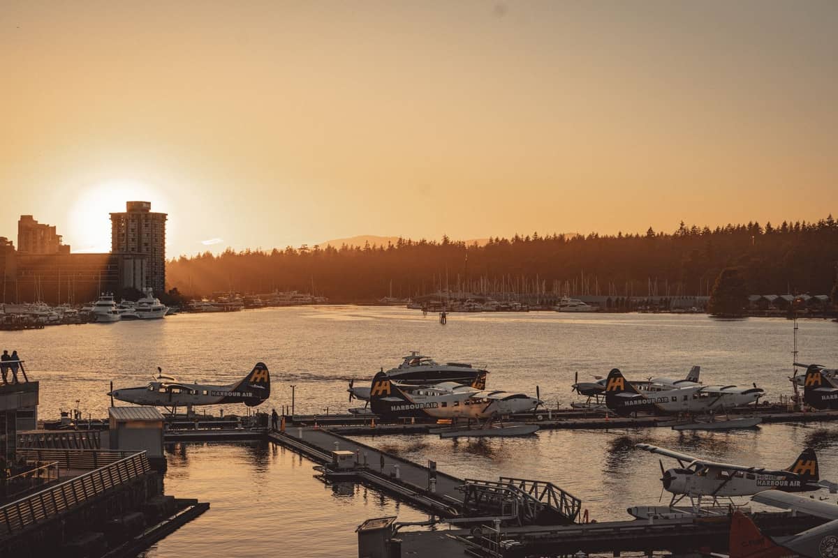 Mehrere Wasserflugzeuge von Harbour Air am Terminal am Ufer von Vancouver bei Sonnenuntergang, mit der Skyline der Stadt und dem Stanley Park im Hintergrund.