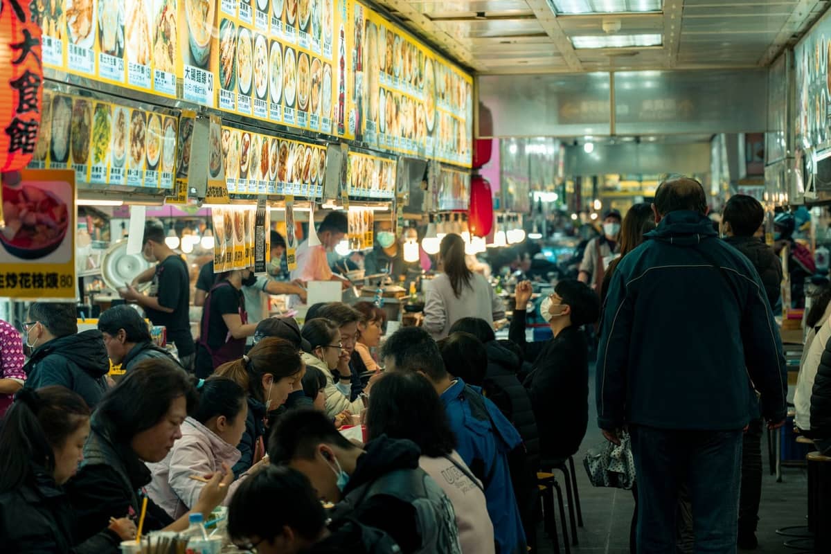 Belebtes Food-Court mit Menschen, die an Gemeinschaftstischen unter hellen Menütafeln speisen und verschiedene asiatische Gerichte in einem lebhaften Innenbereich anbieten.