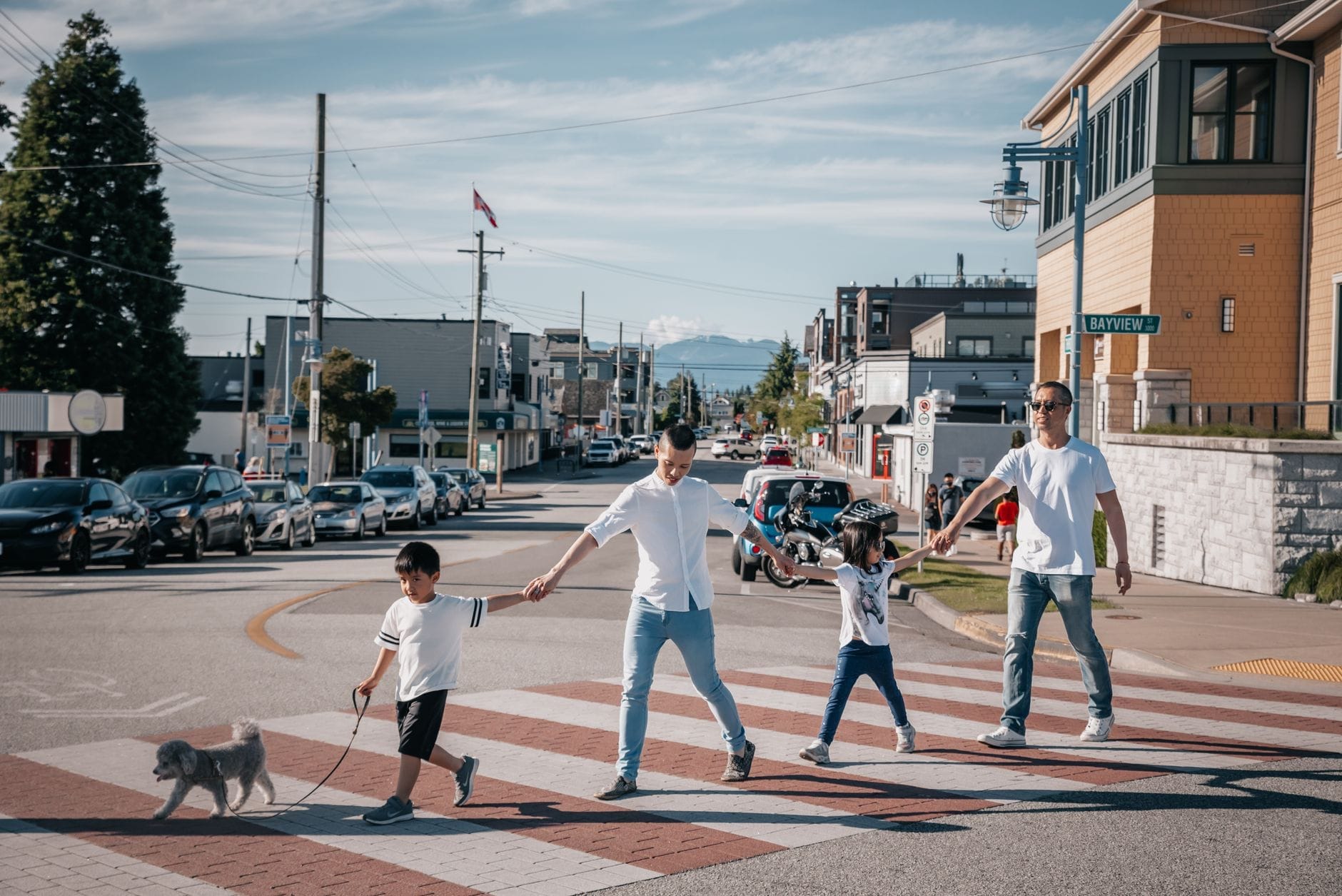 Uma família com crianças pequenas, de mãos dadas e passeando com um cachorro, atravessa uma rua tranquila em um bairro ensolarado e seguro de Vancouver, com casas e carros estacionados ao redor.