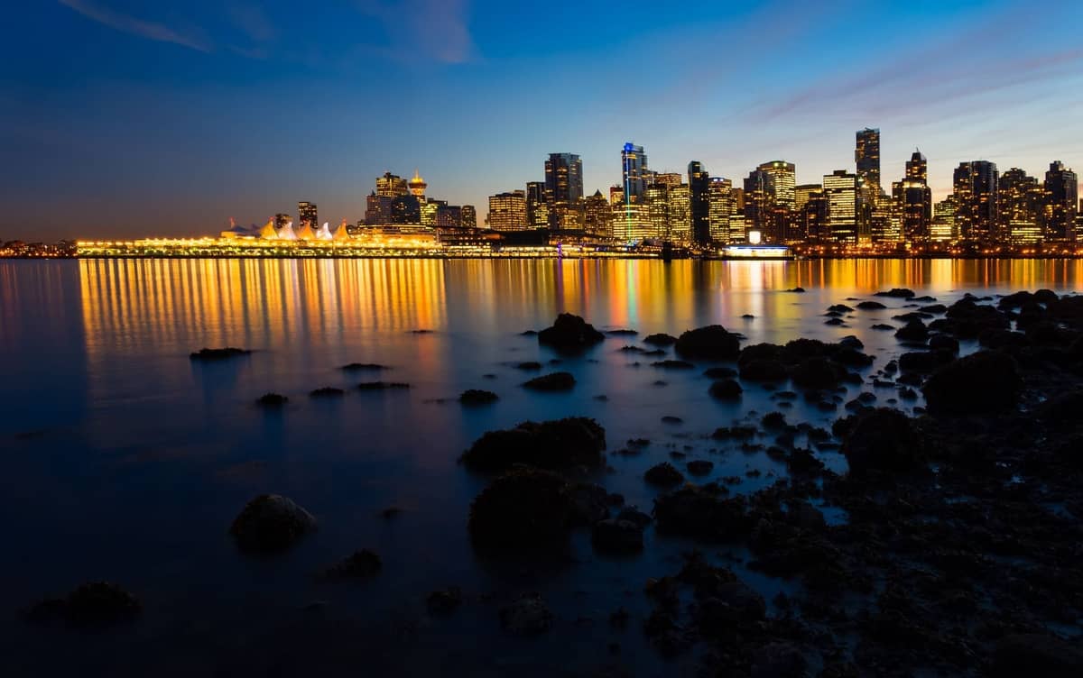 เส้นขอบฟ้าของ Vancouver ยามพลบค่ำ แสงไฟของเมืองสะท้อนบนผืนน้ำพร้อมชายฝั่งหินในเบื้องหน้า