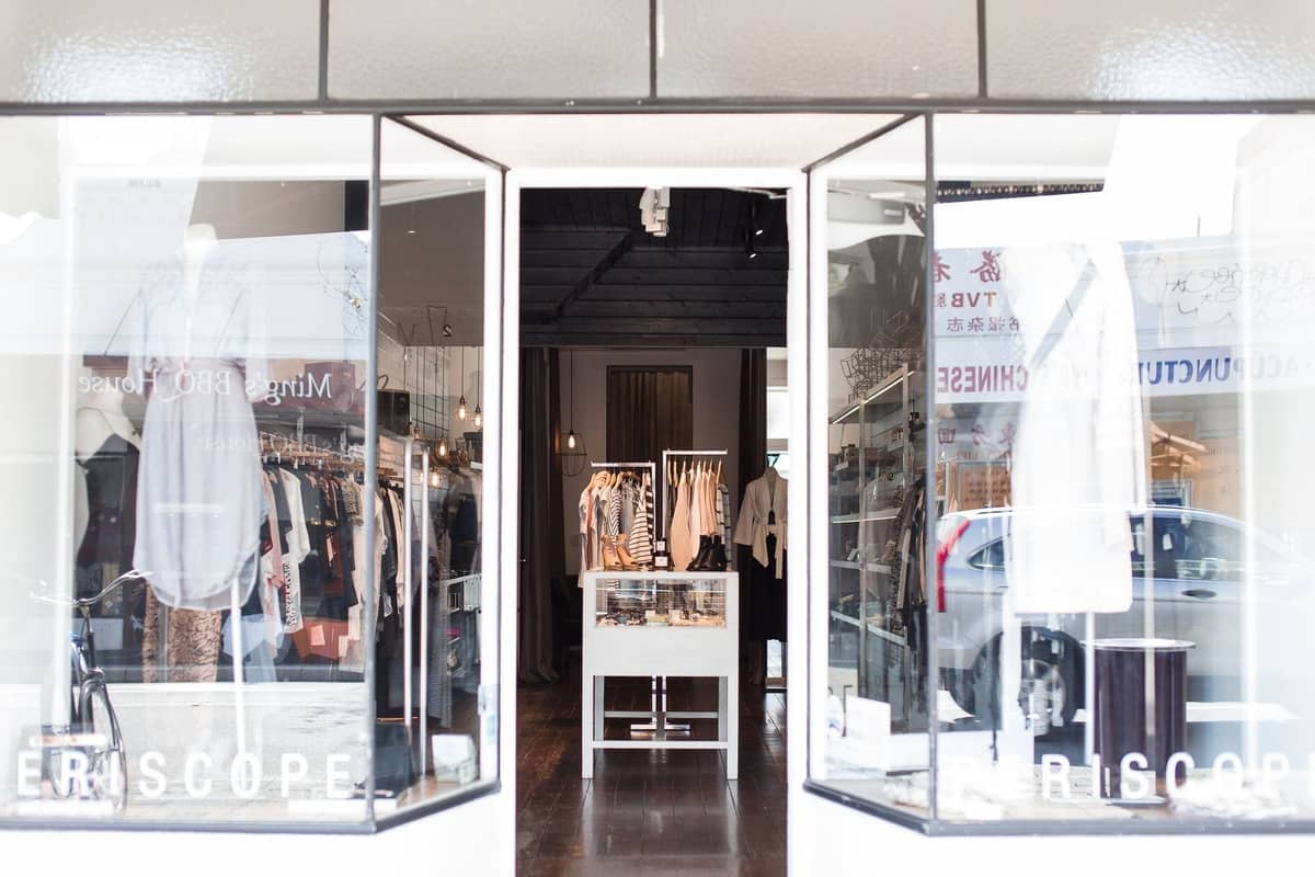 Hell erleuchtete Boutique-Front mit Kleidungsständern und Accessoires, von der Straße aus mit offenen Glastüren gesehen, die Kunden einladen einzutreten.