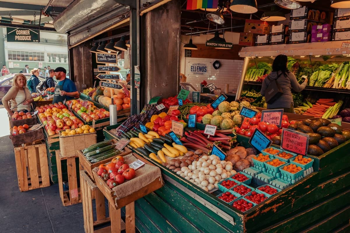 Belebter überdachter Markt mit bunten Obst- und Gemüseständen, Einkäufern und handgeschriebenen Preisschildern, wahrscheinlich der Granville Island Public Market in Vancouver.