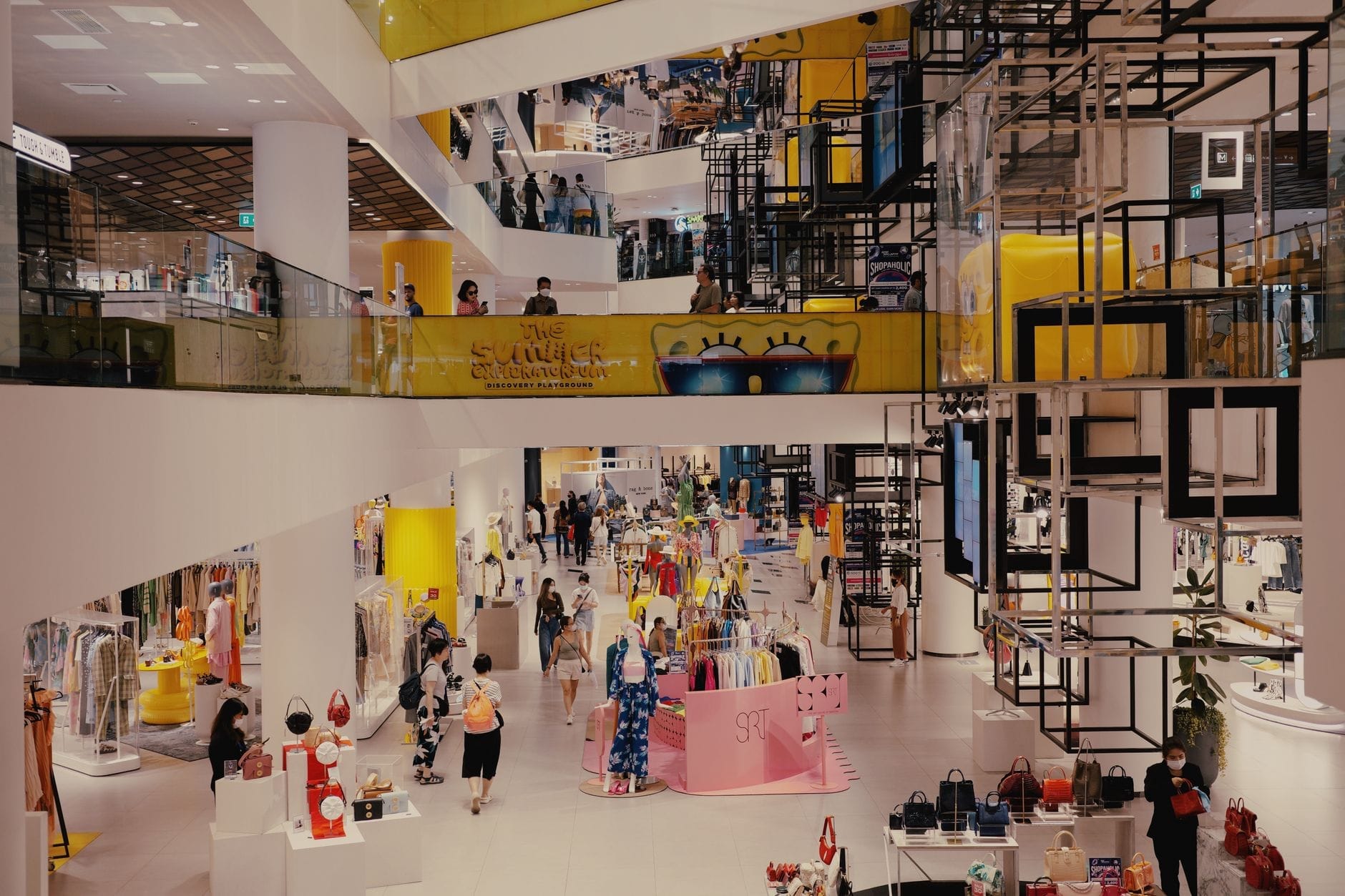 Interior amplo de um shopping moderno com vários andares, visitantes circulando entre vitrines coloridas e uma atmosfera animada típica do varejo urbano.