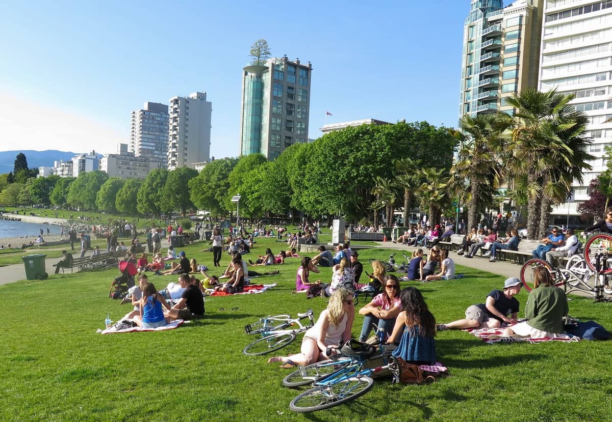Menschen, die an einem sonnigen Tag auf dem Rasen in einem Park am Strand in Vancouver entspannen, mit Stadtgebäuden und Grünflächen im Hintergrund.