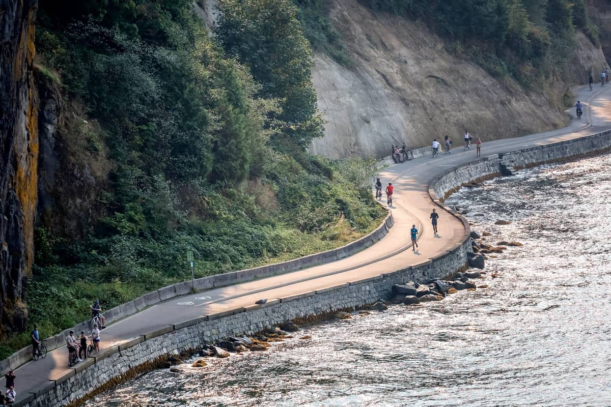 Menschen, die entlang des ikonischen Stanley Park Seawall-Wegs in Vancouver neben dem Ozean und bewaldeten Klippen spazieren und Fahrrad fahren.