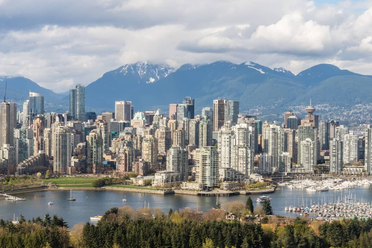 ภาพถ่ายทางอากาศในเวลากลางวันของเส้นขอบฟ้าเมือง Vancouver พร้อมอาคารสูง ท่าเรือ และภูเขาที่ปกคลุมด้วยหิมะในพื้นหลัง