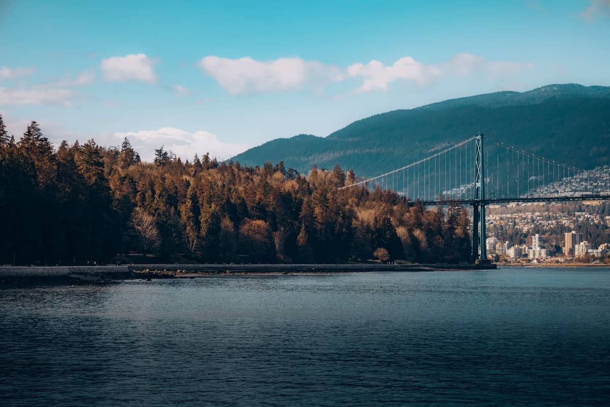 วิวของ Lions Gate Bridge ในเมือง Vancouver พร้อมเส้นขอบฟ้าของตัวเมืองและภูเขาที่ปกคลุมด้วยป่าไม้เป็นฉากหลัง มองจากฝั่งตรงข้ามของน้ำ