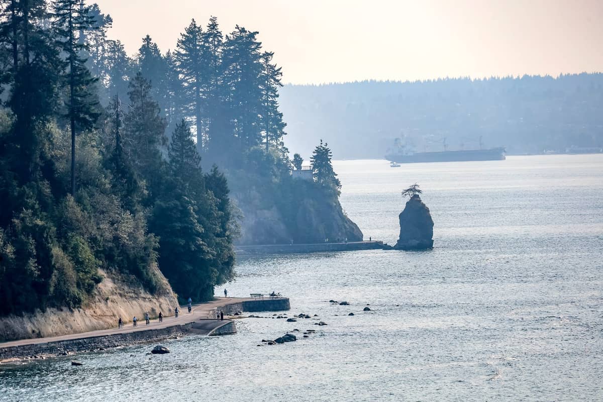 ทางเดิน Seawall ของ Stanley Park ในเมือง Vancouver พร้อมต้นไม้และมหาสมุทร เกาะหินเล็กๆ และเรือในระยะไกลในวันที่แสงสดใส