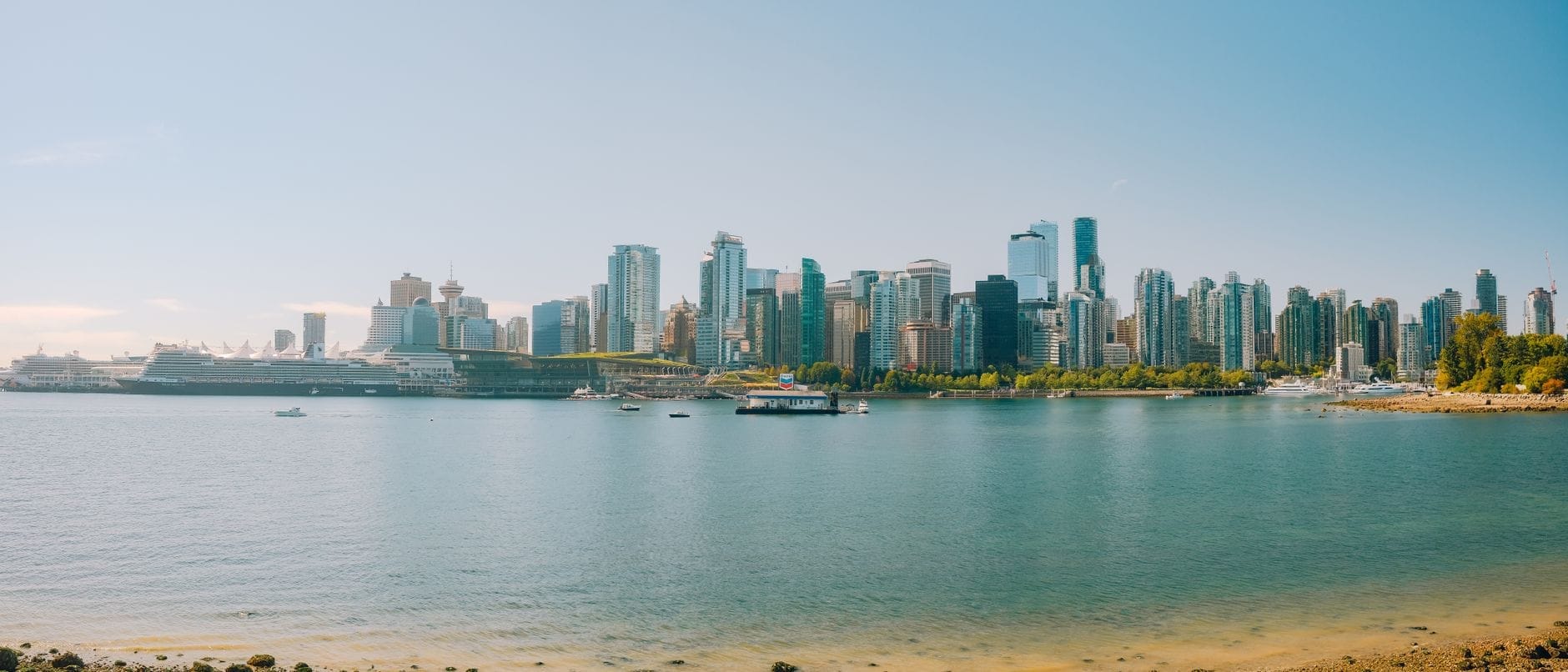Vista panorâmica ampla do skyline de Vancouver em um dia ensolarado, com arranha-céus, água azul em primeiro plano e céu limpo ao fundo.