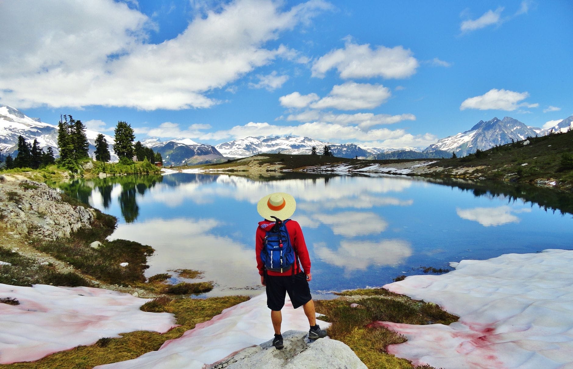 Um caminhante com mochila e chapéu de sol está em uma margem rochosa, contemplando um lago alpino com manchas de neve, cercado por montanhas e céu azul.