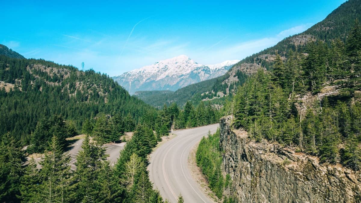 Eine kurvenreiche Bergstraße mit grünen Bäumen und schneebedeckten Gipfeln unter einem blauen Himmel, die ideale Landschaft für einen Sommer-Roadtrip.