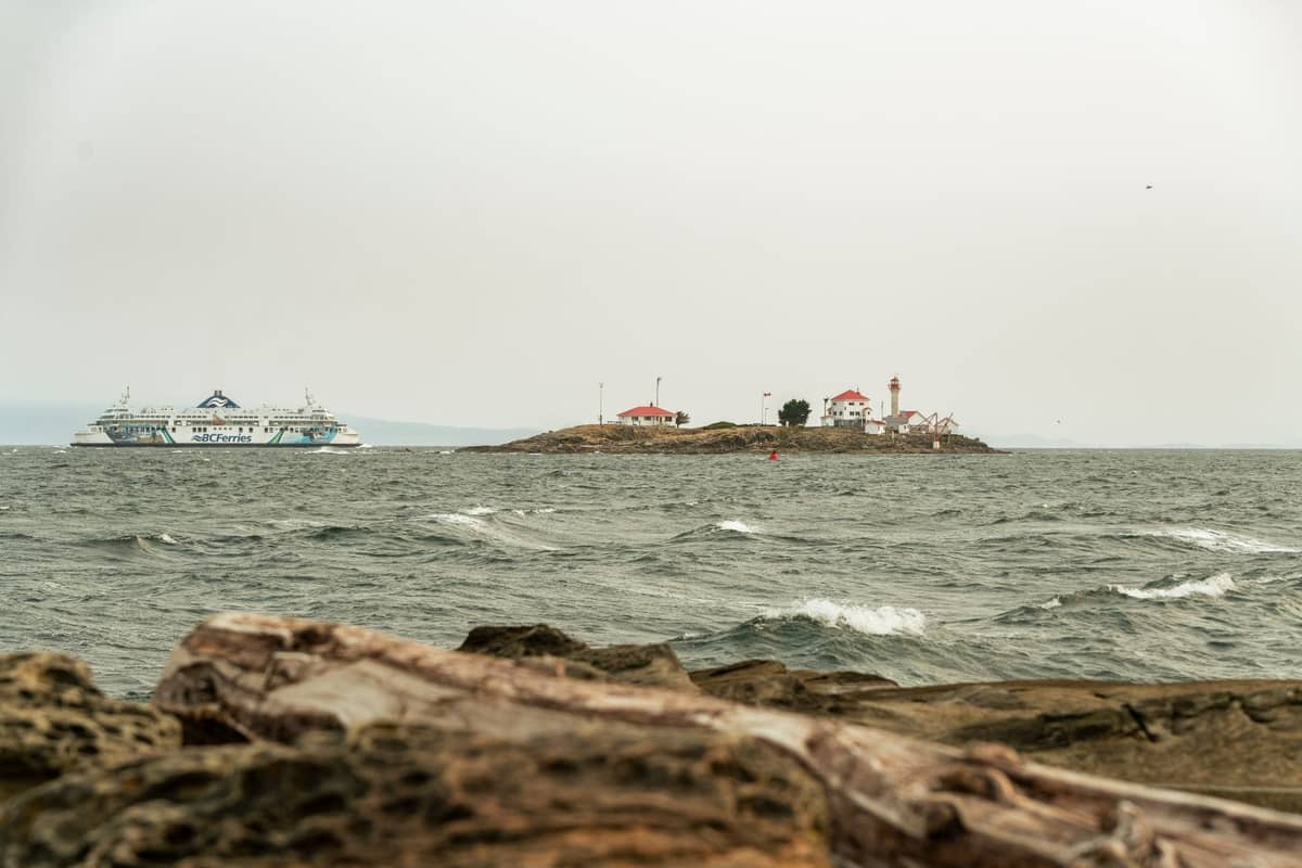 Blick über das Wasser mit einem BC Ferries-Schiff und einem Leuchtturm auf einer kleinen Insel unter bewölktem Himmel, als Symbol für Reisen zwischen Vancouver und Vancouver Island.