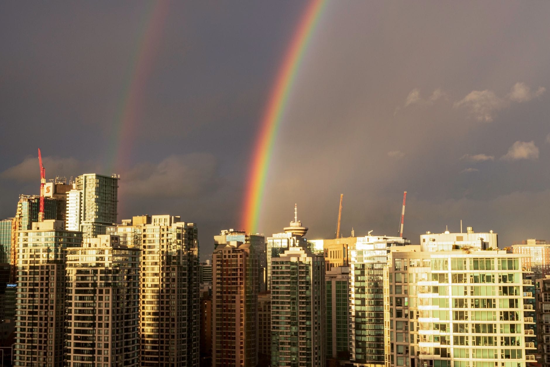 Arco-íris duplo sobre os arranha-céus do centro de Vancouver com nuvens dramáticas, simbolizando o clima variável da cidade e sua paisagem urbana.