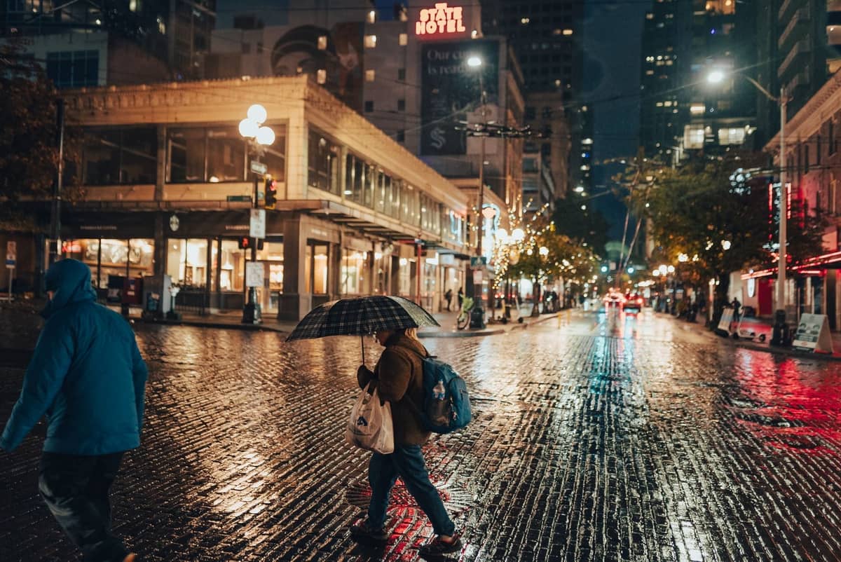 Regnerische Abendstraßenszene in der Innenstadt von Vancouver mit Menschen, die unter Regenschirmen gehen, und Spiegelungen auf dem nassen Pflaster.