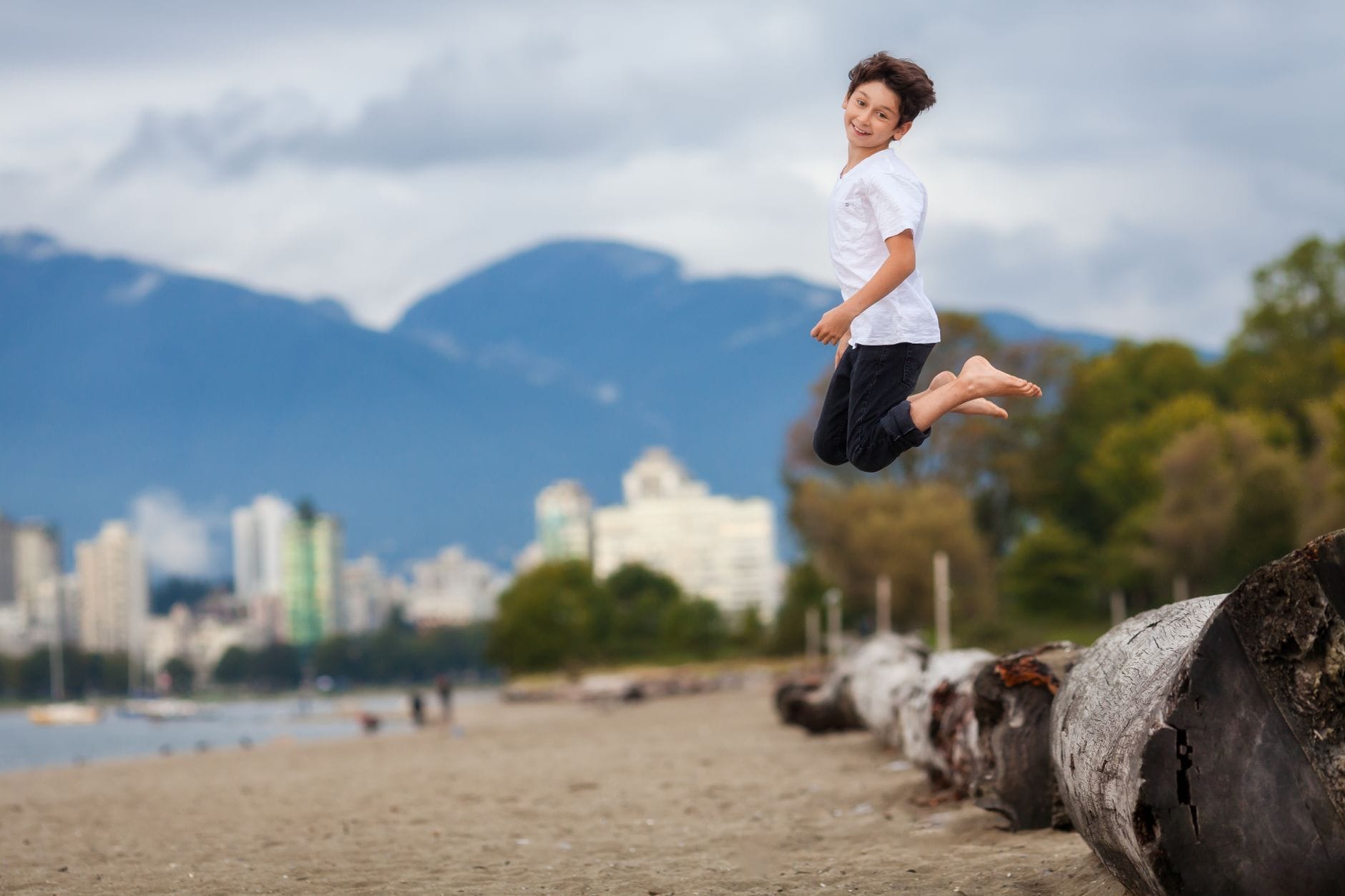Um menino feliz pula alto em uma praia de areia em Vancouver com arranha-céus e montanhas ao fundo, evocando diversão e aventura em família.
