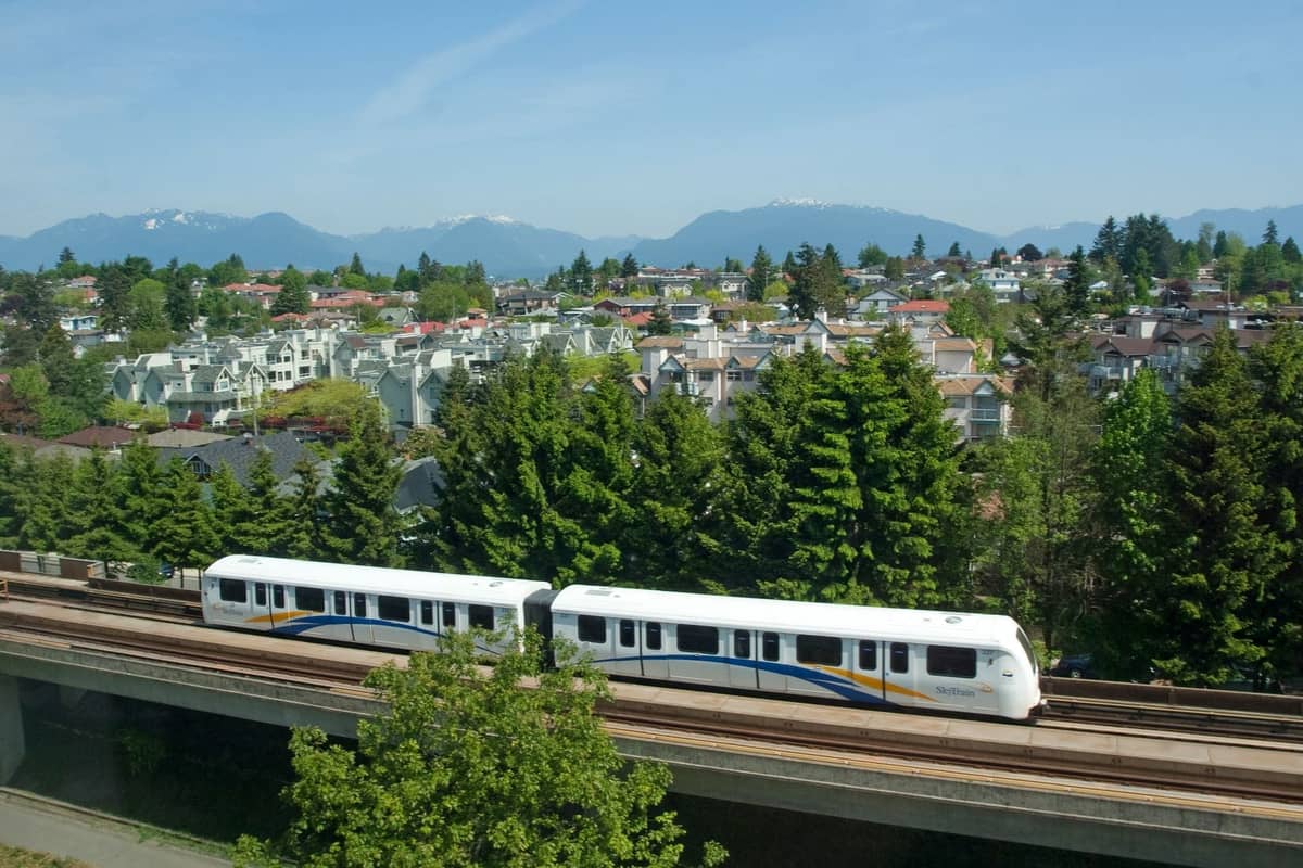 Ein Vancouver SkyTrain gleitet auf Hochgleisen über grüne Wohnviertel, mit im Hintergrund sichtbaren Bergen unter einem klaren Himmel.