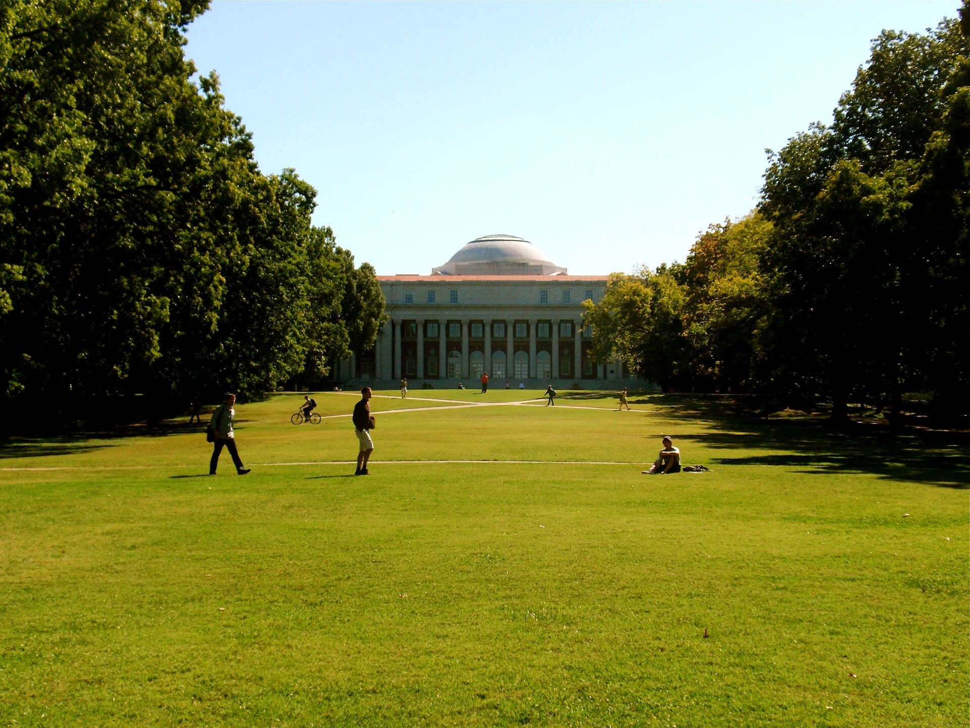 Amplio césped en el campus de Vanderbilt University con personas caminando y en bicicleta bajo cielos soleados, enmarcado por grandes árboles y un histórico edificio con cúpula.