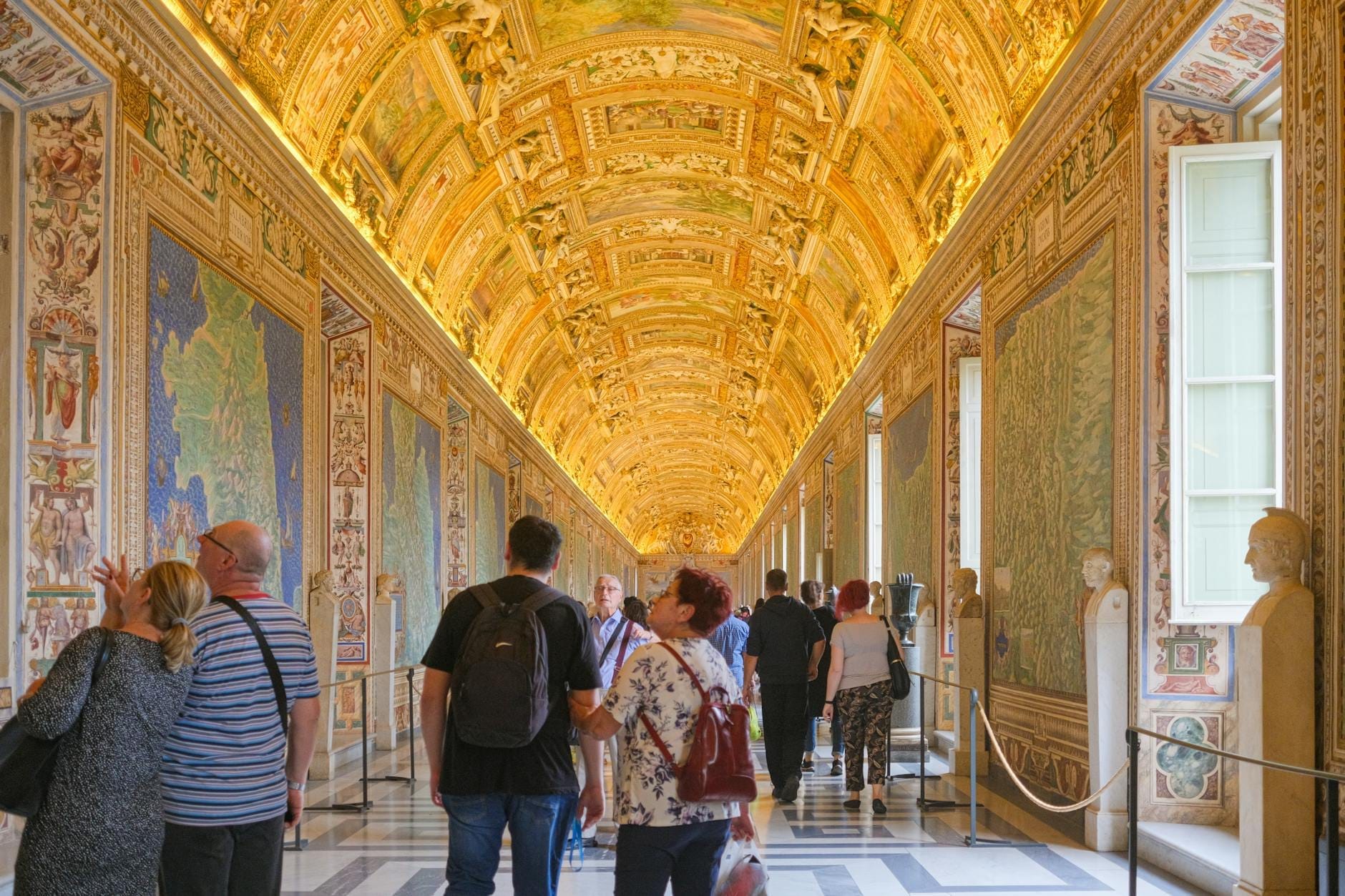 Visitors walking through the ornately decorated Gallery of Maps in the Vatican Museums, with golden vaulted ceilings and richly colored frescoes.