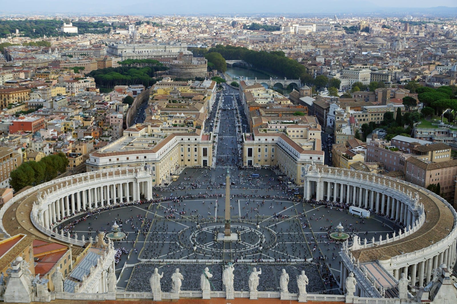 A panoramic view of St. Peter's Square with its iconic colonnades, statues, and cityscape of Rome in the background under a clear sky.
