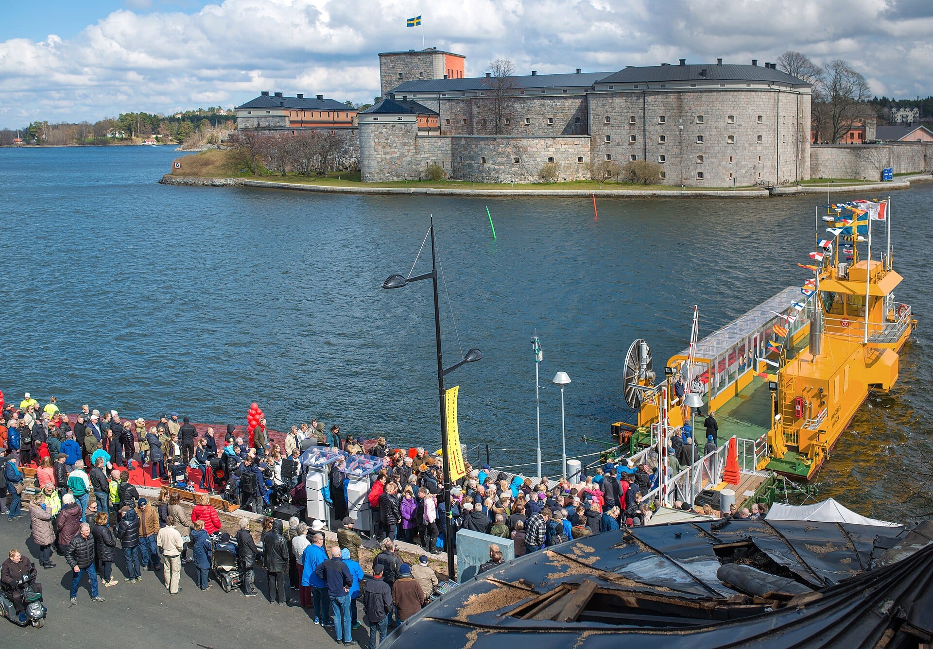 ป้อมปราการ Vaxholm มองจากมุมสูง เห็นเรือเฟอร์รี่สีเหลืองจอด มีคนพลุกพล่าน ริมน้ำและเมืองอยู่ด้านหลัง