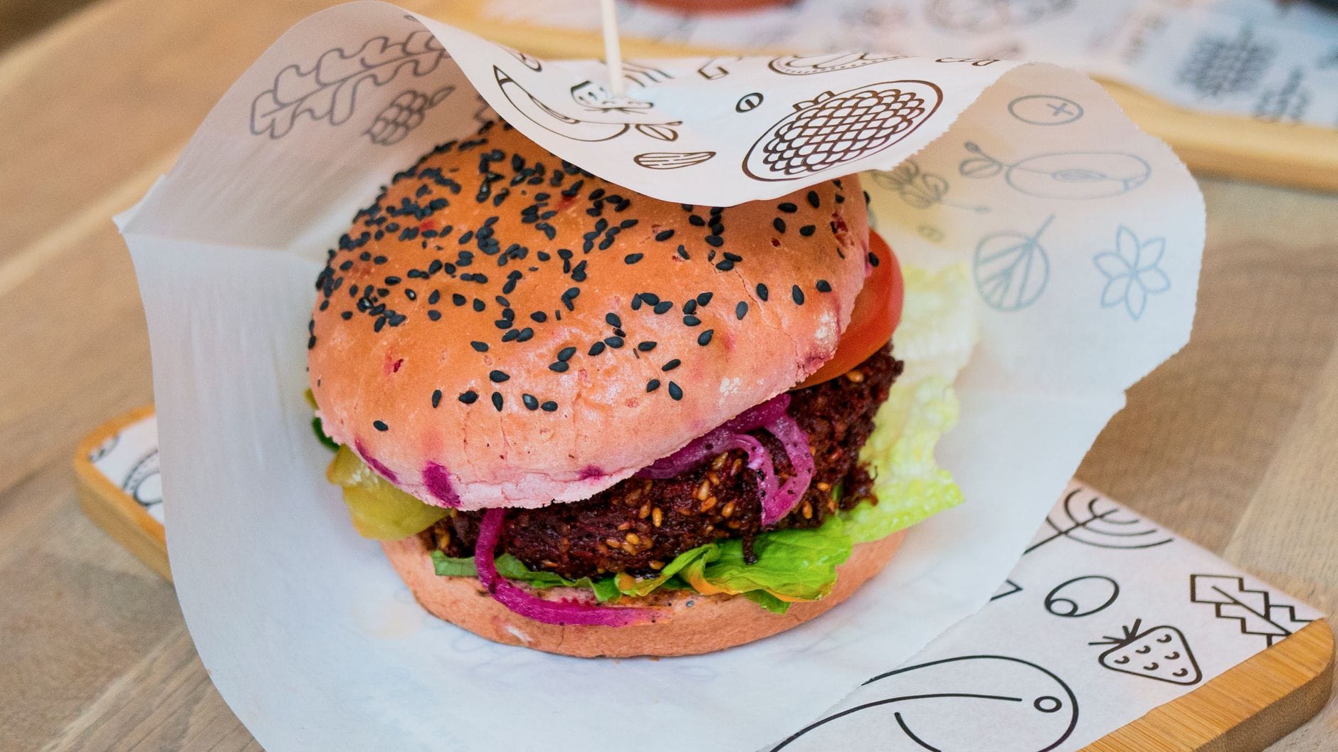 A colorful vegan burger with a pink bun, lettuce, tomato, and patty, served on decorative paper in a bright, inviting setting.