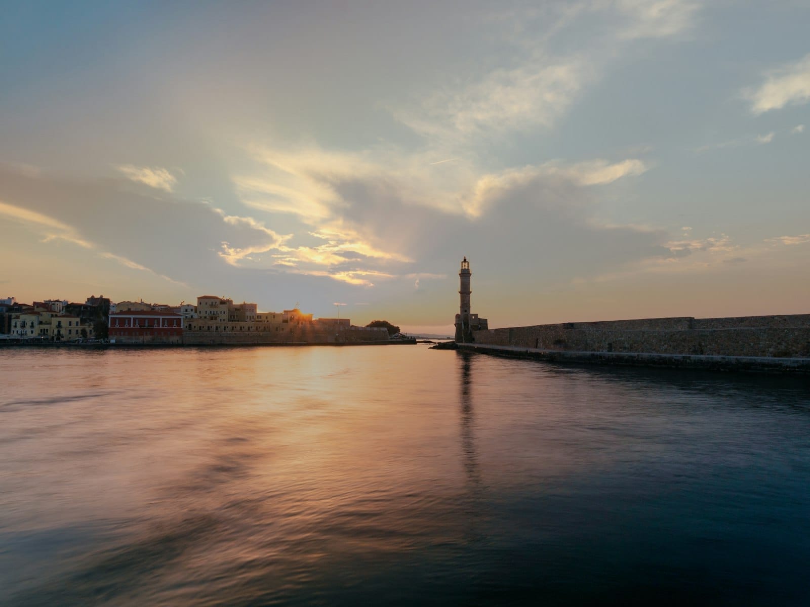 Tramonto sul Porto Veneziano di Canea con l'iconico faro e gli edifici storici del lungomare riflessi in un'acqua calma e colorata sotto un cielo drammatico.