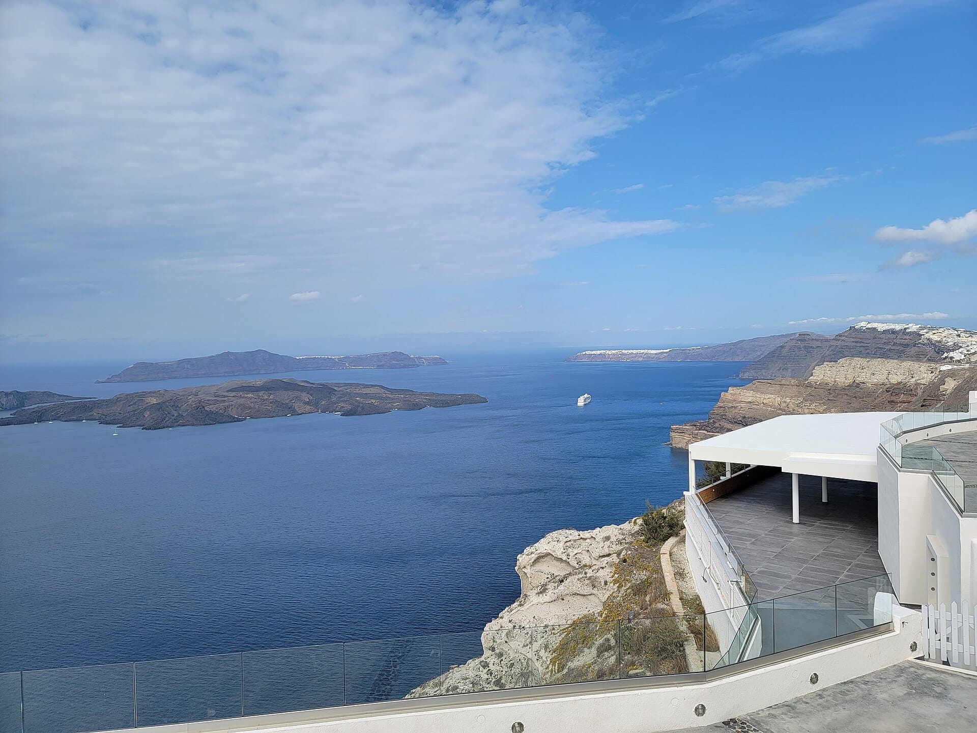 ระเบียงของ Venetsanos Winery มองเห็นปากปล่องภูเขาไฟสีฟ้าของ Santorini หน้าผาสูงชัน เกาะที่อยู่ไกลออกไป และเรือสำราญสีขาวในวันที่แดดจ้า