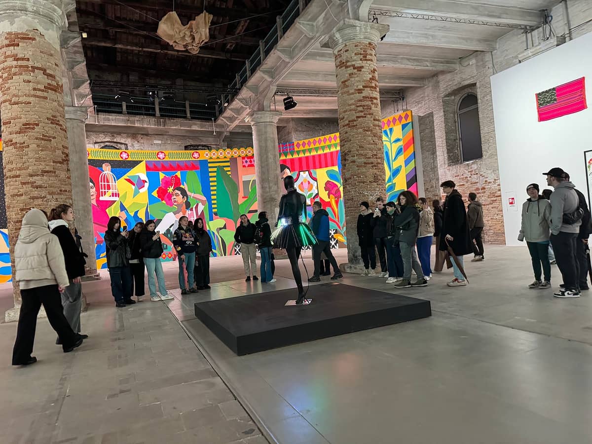 Besucher erkunden eine zeitgenössische Kunstinstallation in der Arsenale-Halle der Venedig Biennale, umgeben von leuchtenden Wandgemälden und historischen Backsteinpfeilern.