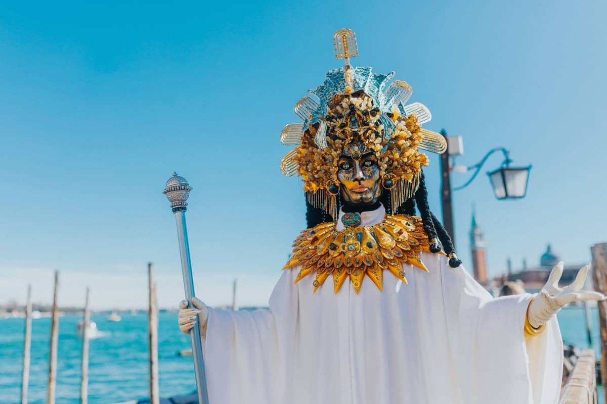 Eine Person in einem aufwendigen gold-weißen venezianischen Karnevalskostüm mit Maske steht am Wasser, im Hintergrund die Skyline Venedigs und ein blauer Himmel.