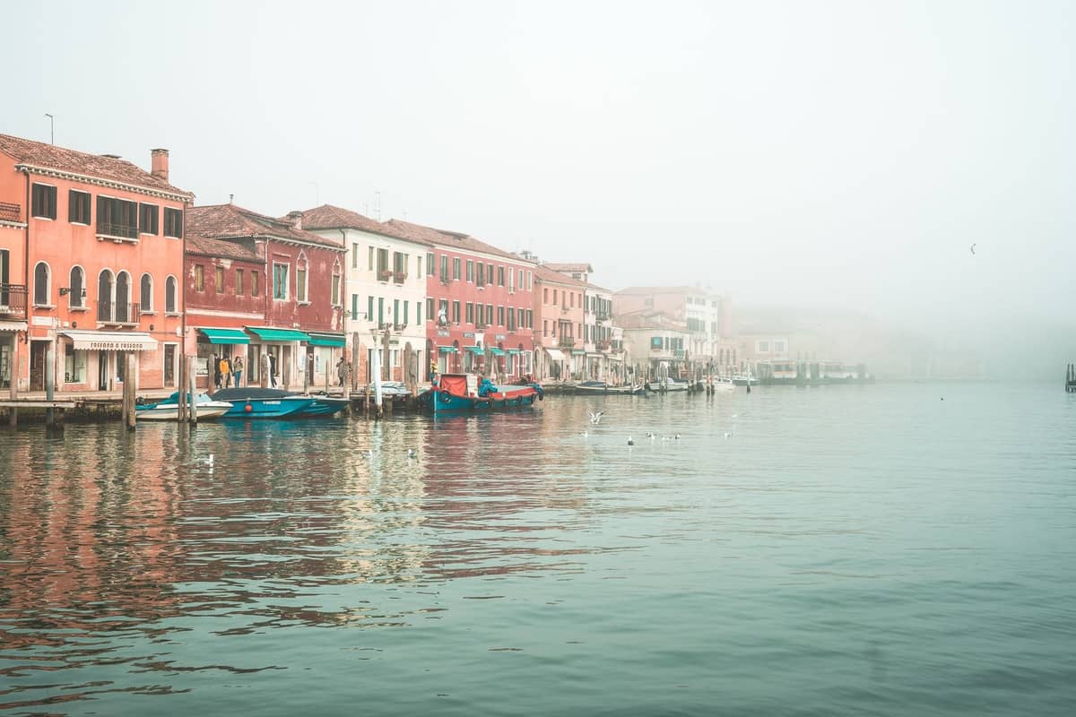 Ein ruhiger, nebeliger venezianischer Kanal, gesäumt von pastellfarbenen Wohnhäusern mit einigen Booten und ohne Menschenmassen, der die lokalen Viertel Venedigs widerspiegelt.