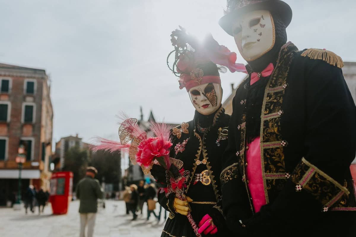 Zwei Personen in aufwendigen, traditionellen venezianischen Karnevalskostümen und Masken, die im Freien auf einem venezianischen Platz stehen.