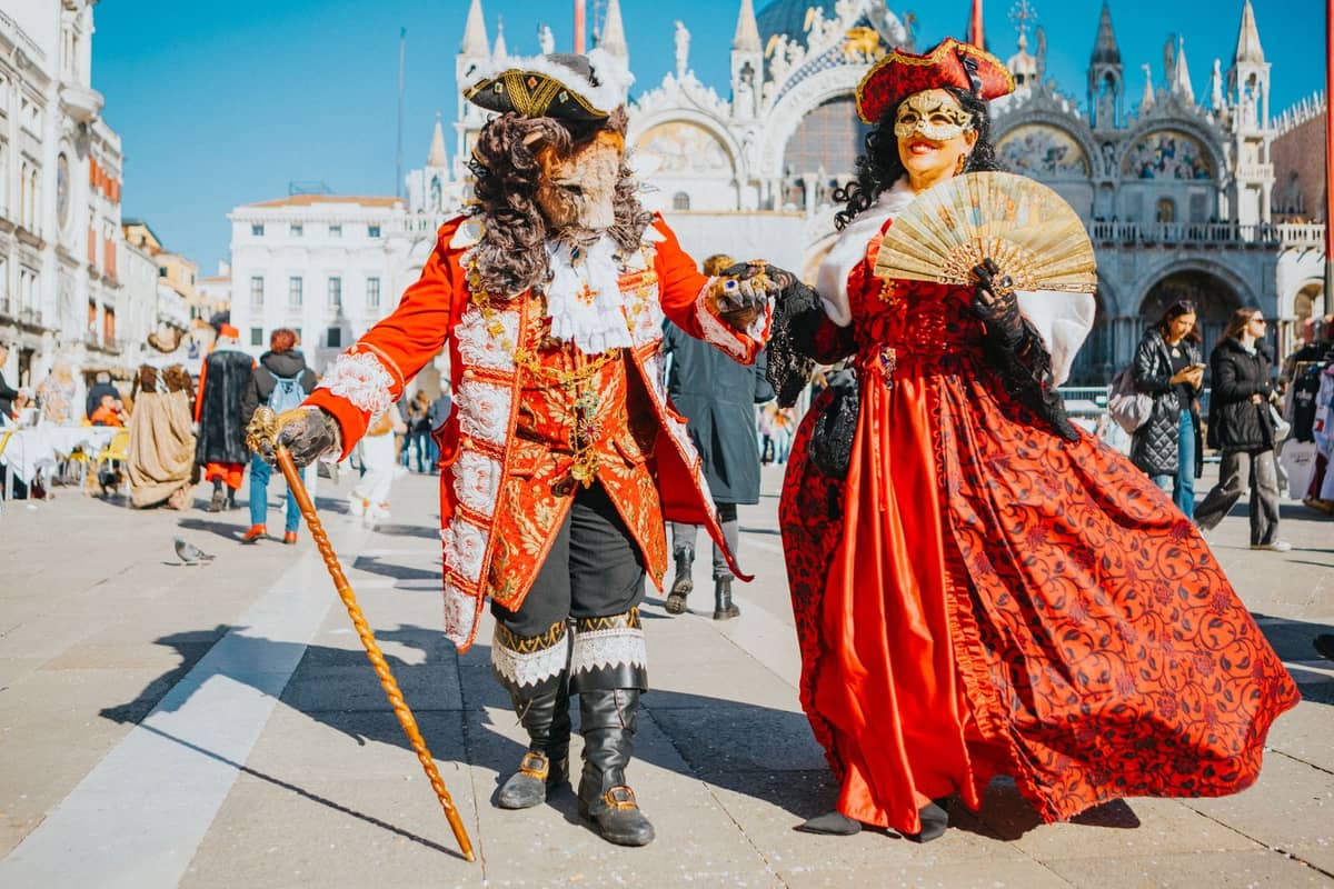 Zwei Personen in aufwendigen historischen venezianischen Karnevalskostümen und Masken stehen auf der Piazza San Marco, mit der Basilica di San Marco im Hintergrund.