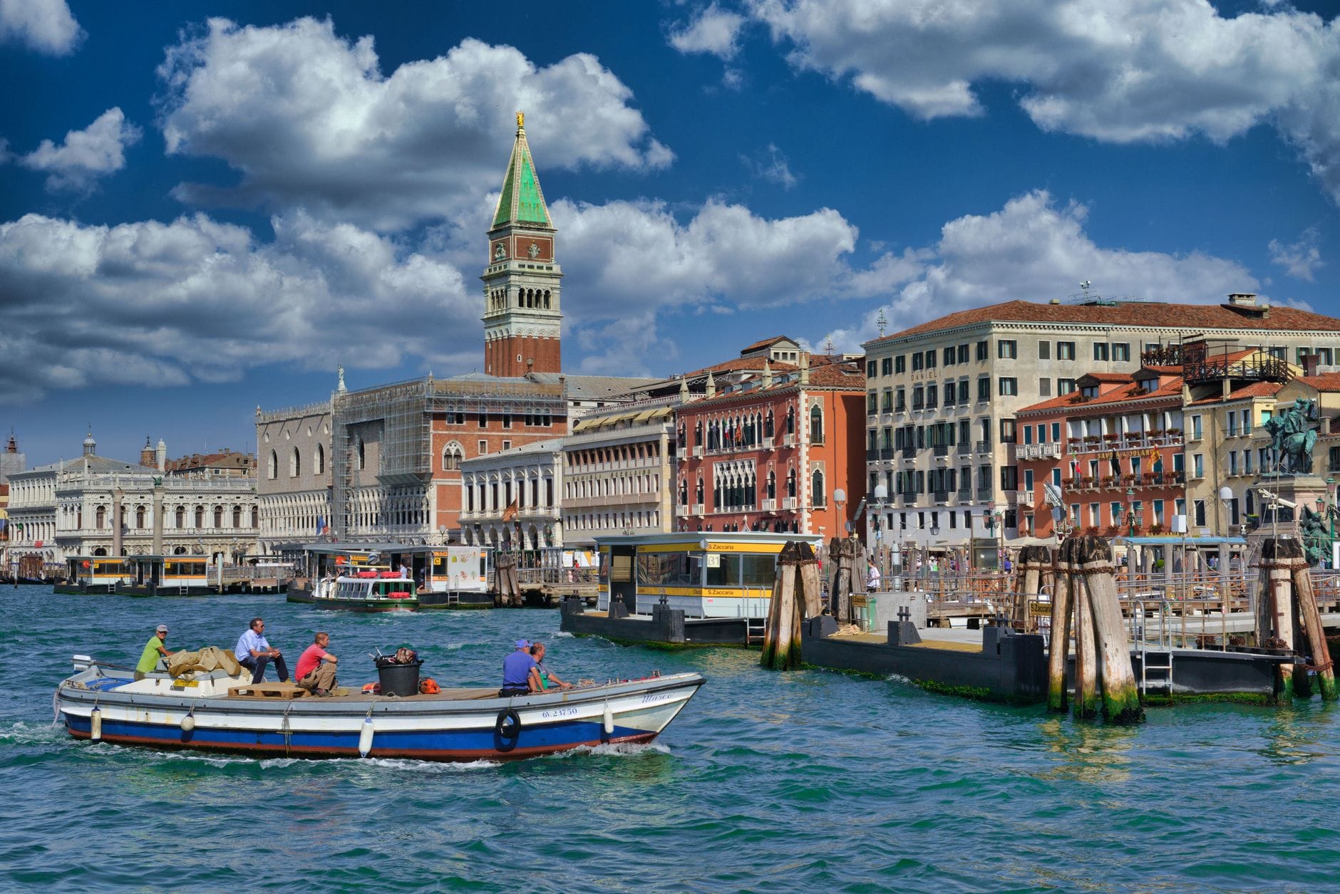 Classic view of Venice featuring the Grand Canal with boats, colorful historic buildings, and the iconic St Mark’s Campanile under a bright blue sky.