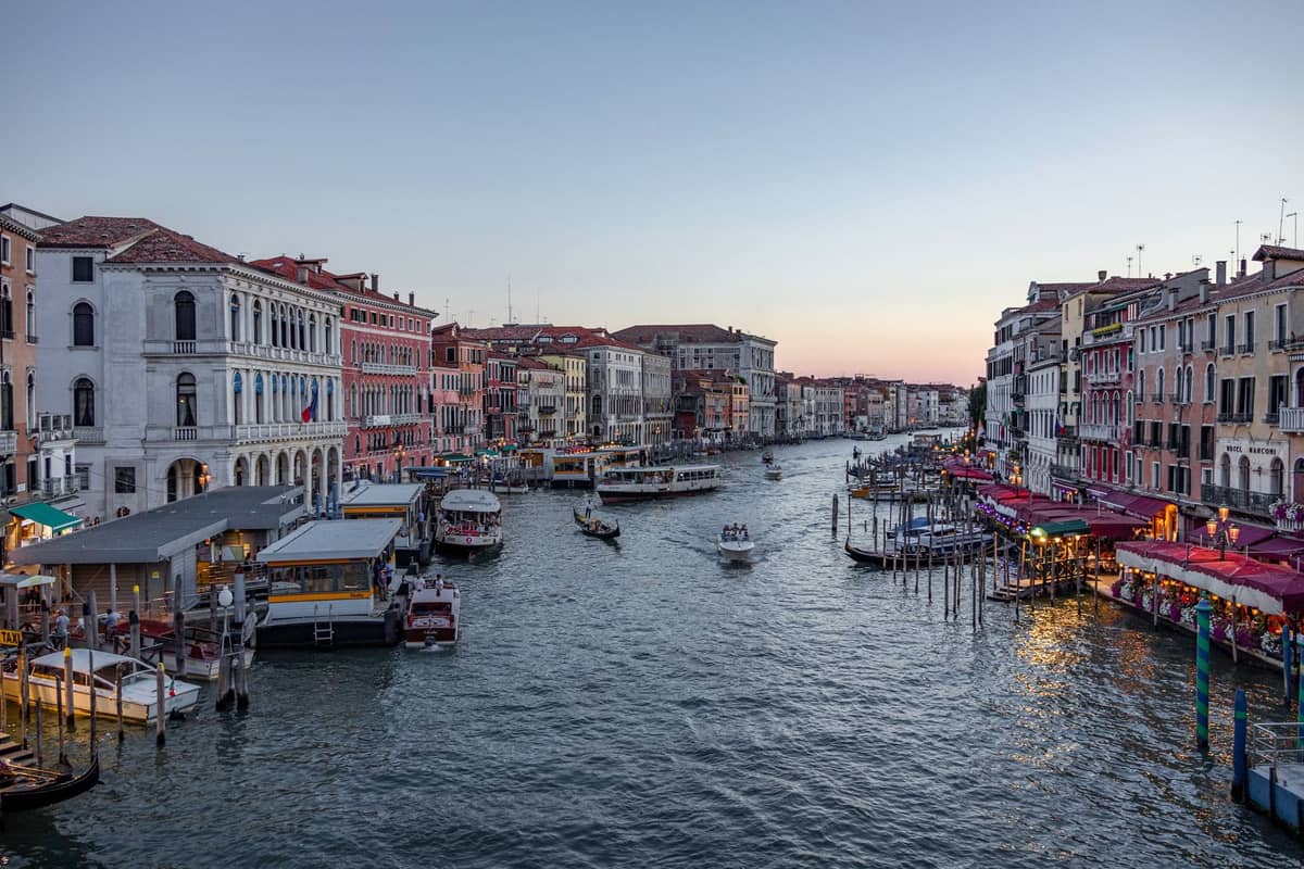 Blick den Canal Grande in Venedig bei Sonnenuntergang hinunter mit belebten Uferhotels, Cafés, Booten und Vaporetto-Haltestellen auf beiden Seiten des Kanals.