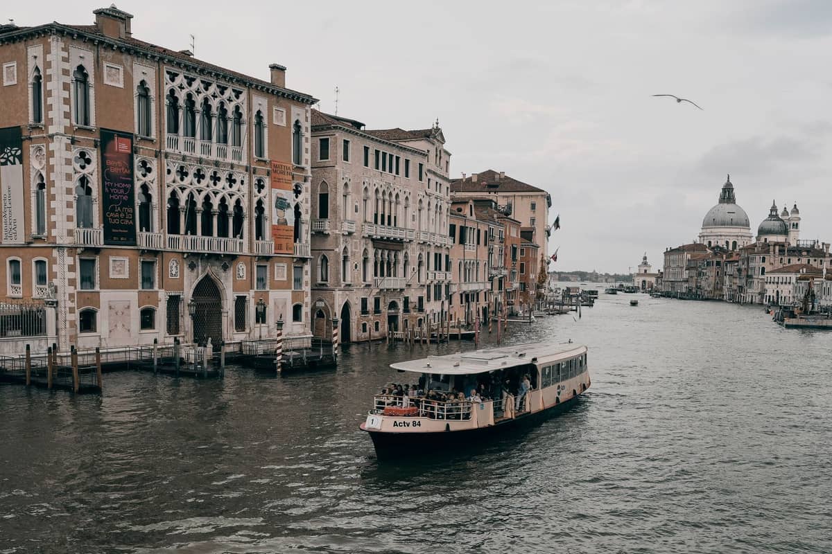 Vaporetto fährt den Canal Grande in Venedig entlang mit historischen Gebäuden und Kuppeln im Hintergrund unter bewölktem Himmel.