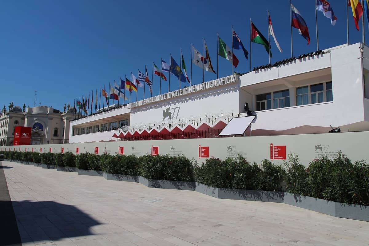 Weitwinkelperspektive auf die Hauptfassade des Palazzo del Cinema, geschmückt mit internationalen Flaggen und Festivalbannern unter strahlend blauem Himmel auf dem Lido di Venezia.