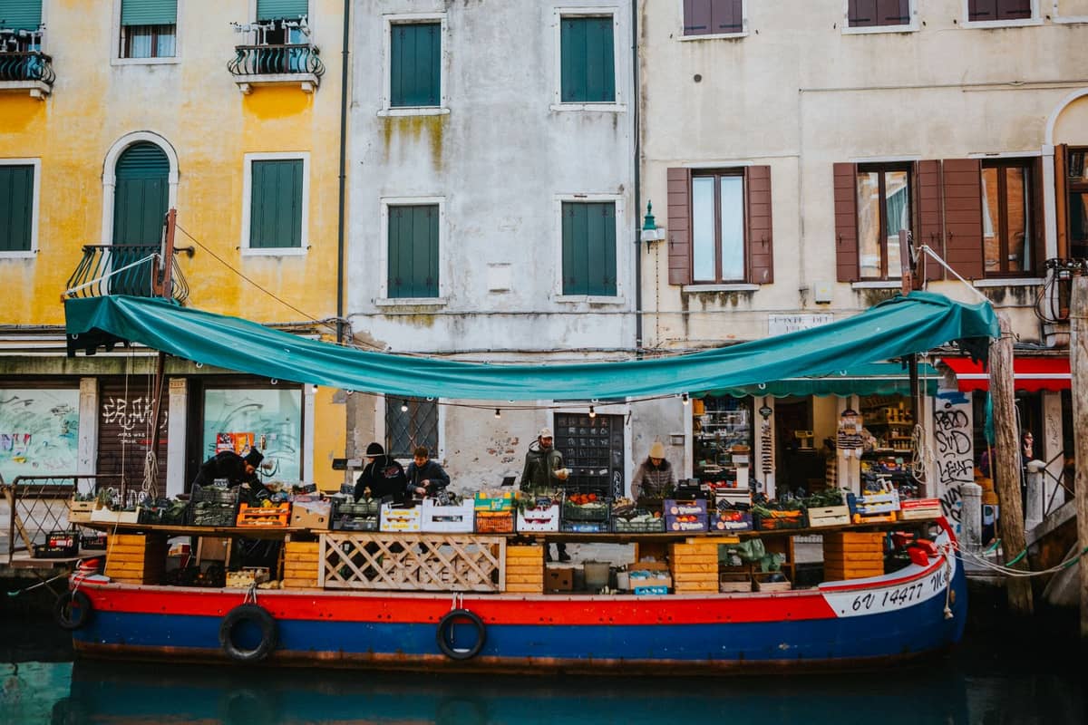 Buntes schwimmendes Marktboot mit Produkten auf einem Kanal in Venedig mit lebhaften Gebäuden im Hintergrund