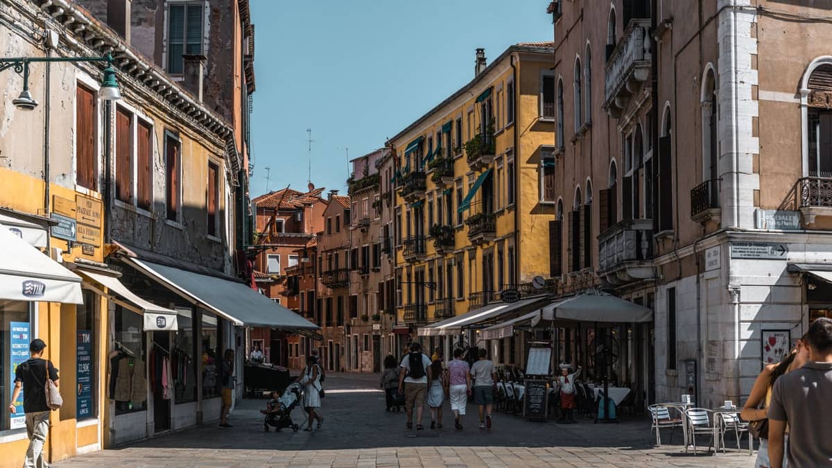 Fußgänger gehen durch eine sonnige, enge venezianische Gasse, gesäumt von historischen Gebäuden und Ladenmarkisen, die die sommerliche Hitze Venedigs heraufbeschwören.