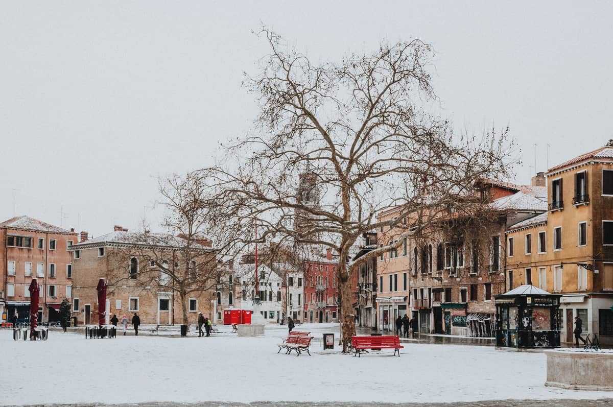 Ein weitläufiger venezianischer Platz mit Schnee, kahlen Bäumen und roten Bänken, umgeben von bunten Wintergebäuden und wenigen Spaziergängern.